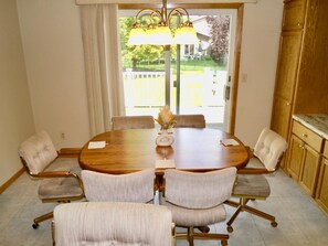 Une salle à manger avec une table ronde, six chaises, un vase de fleurs séchées et une vue sur le jardin à travers une porte vitrée.