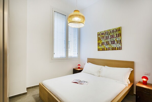 A bedroom with a bed, a bedside table, a nightstand, a window with blinds, and a wall-mounted artwork. - Carignano Sweet Home by Wonderful Italy (Genoa)