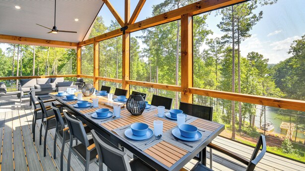 A covered outdoor dining area with a table set for six, surrounded by chairs, and overlooking a forested area with a body of water. - Hot Tub + Game Room! Family Connelly Springs Home (Connellys Springs)