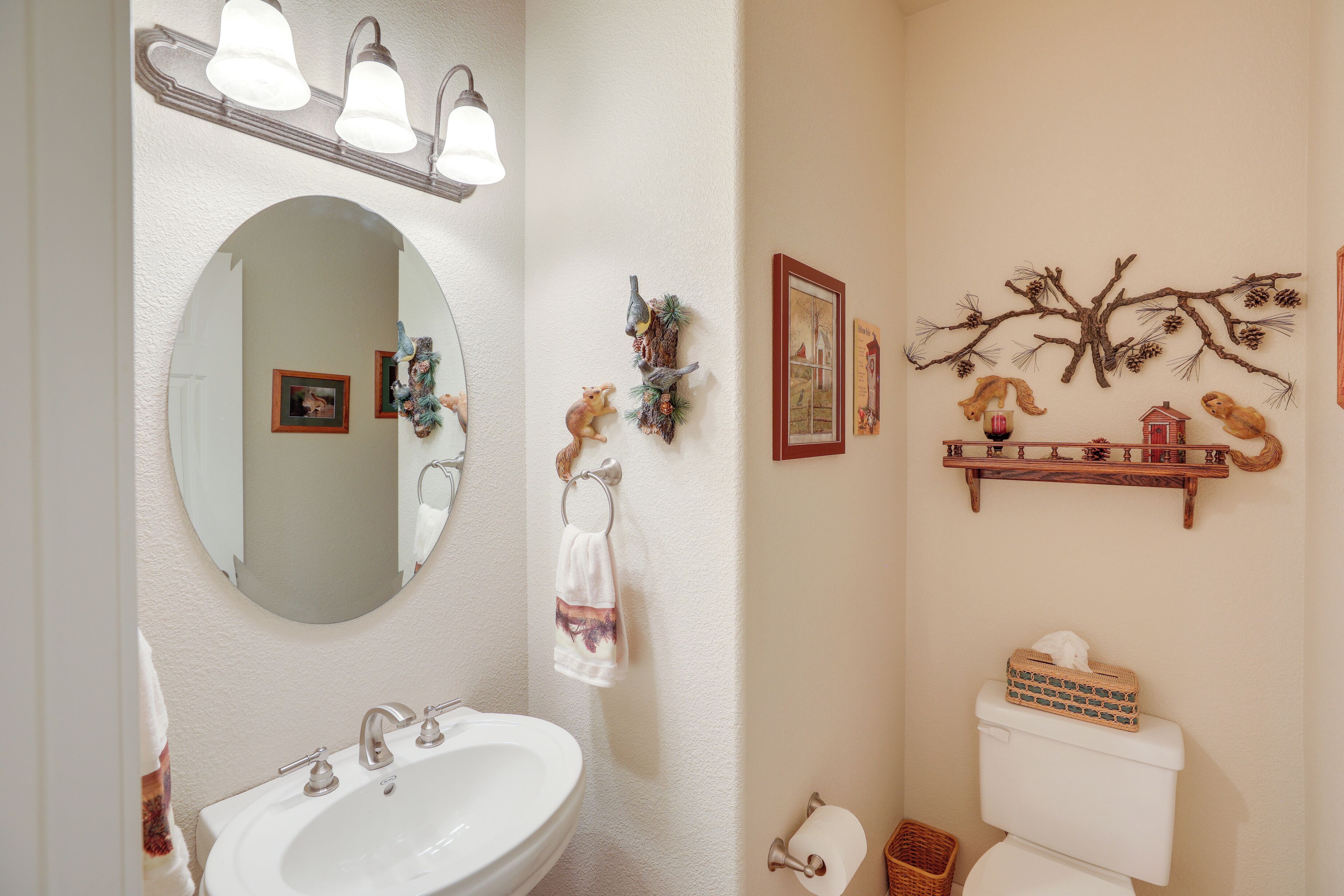 Un baño con un lavabo blanco, un espejo y un inodoro. Se observan adornos en la pared y una repisa con objetos pequeños.