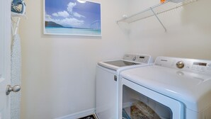 A laundry room with a Samsung washer and dryer, a painting of a beach scene, and a white door.