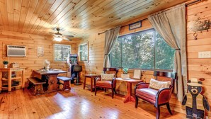 A cozy cabin interior with wooden walls, a dining area, a fireplace, and a large window with curtains. - Bryson City Cabin in Smoky Mtn w/ Resort Amenities (Bryson City)