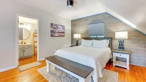 A bedroom with a wooden ceiling, a bed with white bedding, a bench, a nightstand with lamps, and a bathroom visible through an open door. - Lakefront Murphy Getaway w/ Deck, Dock & Kayaks! (Murphy)