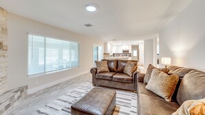 A spacious living room with a leather sofa, a matching ottoman, and a patterned rug. A kitchen area is visible in the background.