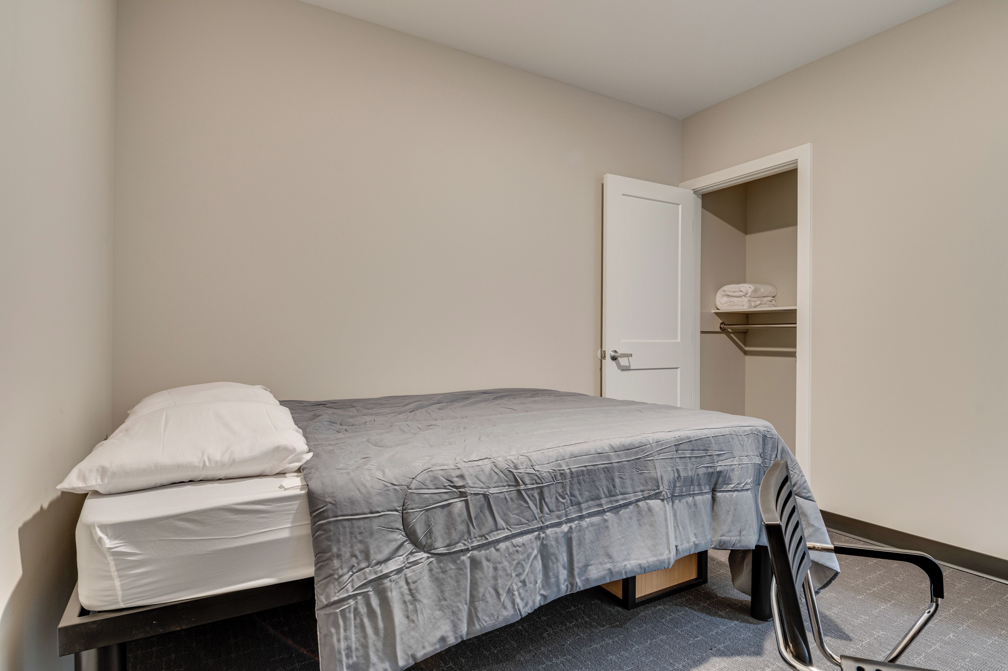 Una cama individual con colchón gris y almohadas blancas, una silla y una puerta que da a un armario con estantes.