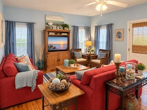 A living room with a red sofa, a wooden coffee table with decorative items, a television on a wooden cabinet, wicker chairs, and a ceiling fan. - Sandy Pines Cottage | Private 3BR, 2 Pools + Beach Nearby (Gulf Shores)