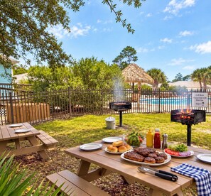 An outdoor barbecue setup with a grill, picnic table, and pool in the background. - Sandy Pines Cottage | Private 3BR, 2 Pools + Beach Nearby (Gulf Shores)