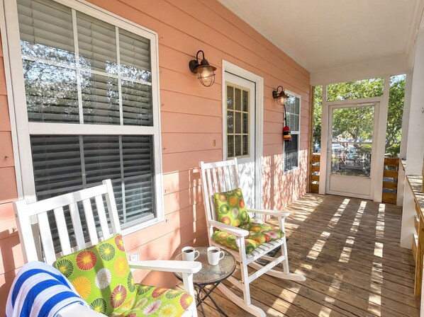 A covered porch with white rocking chairs, a small table with cups, and a door leading to the interior. - Sandy Pines Cottage | Private 3BR, 2 Pools + Beach Nearby (Gulf Shores)