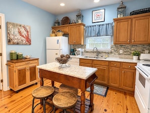 A kitchen with wooden cabinets, a white refrigerator, a granite countertop, and a dining area with stools. - Sandy Pines Cottage | Private 3BR, 2 Pools + Beach Nearby (Gulf Shores)