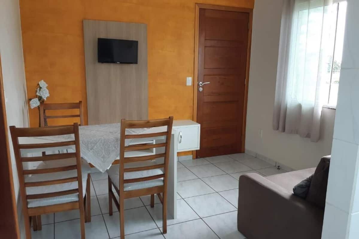 Een eetgedeelte met een tafel en stoelen, een aan de muur bevestigde televisie, een houten deur en een raam met gordijnen.
