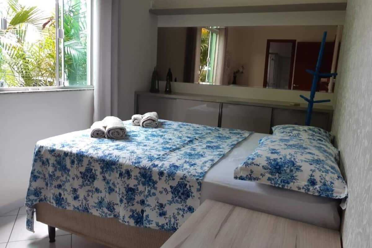 Een slaapkamer met een bed, nachtkastje, spiegel en raam met uitzicht op groen.