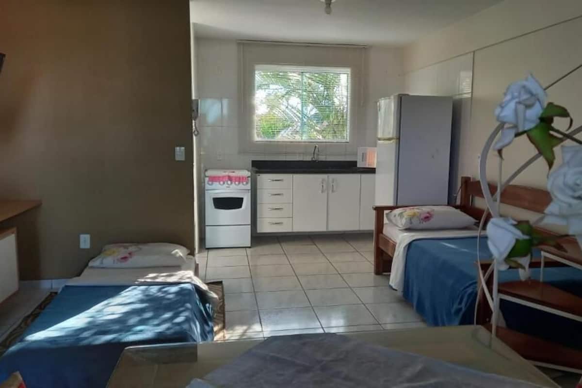 Een kleine, goed verlichte kamer met twee bedden, een kitchenette en een raam met uitzicht op bomen.