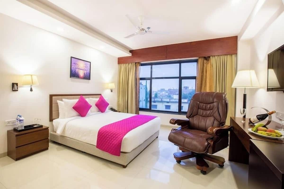 A hotel room with a bed, a desk, a chair, a nightstand, a TV, and a window with curtains.