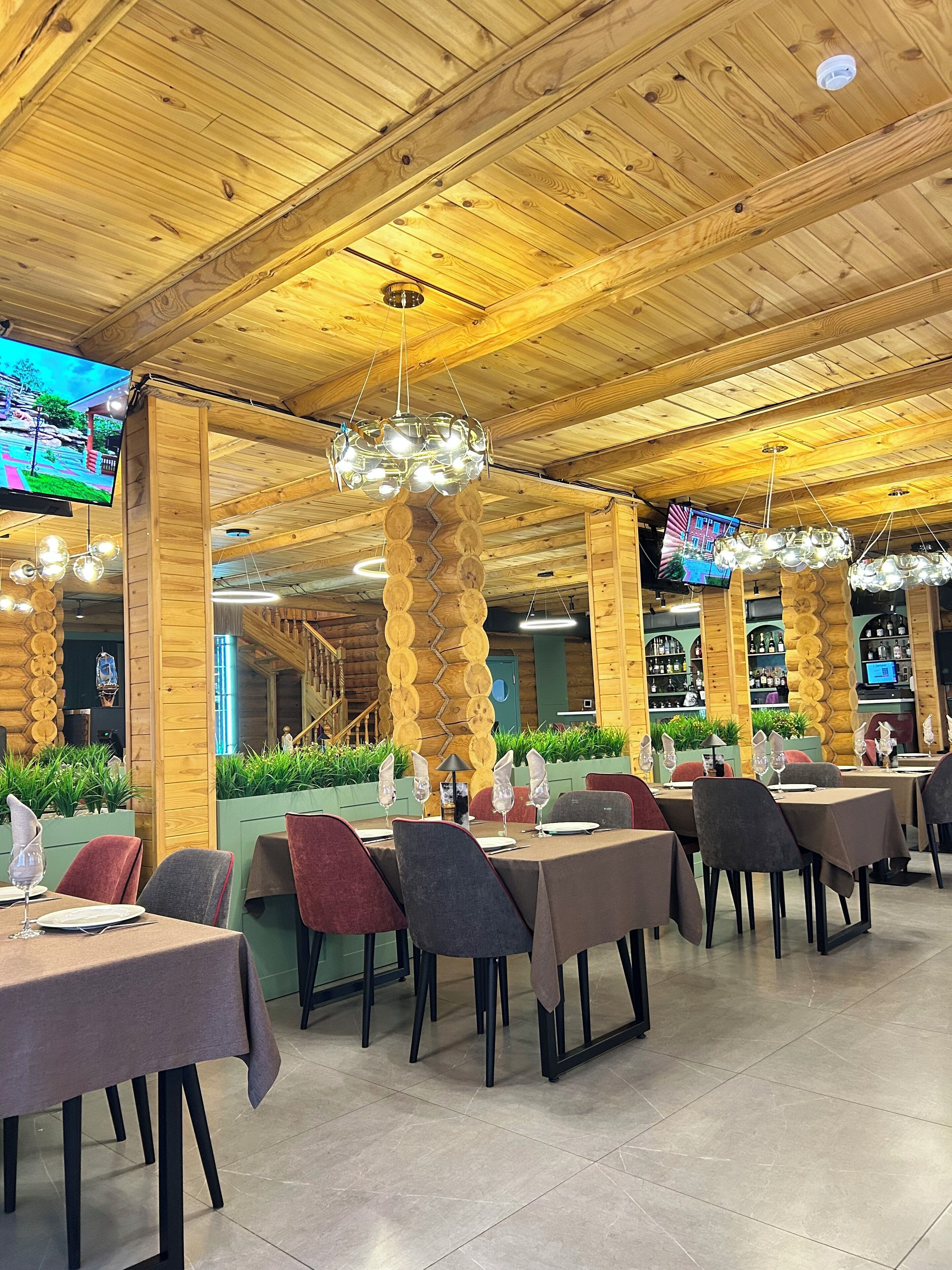 Un restaurante con vigas de madera en el techo, lámparas colgantes y varias pantallas de televisión. Las mesas están dispuestas con sillas y vajilla.