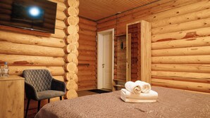 A log cabin room with a bed, a chair, a TV mounted on the wall, and a door leading to another room. - Skala (Burabay)