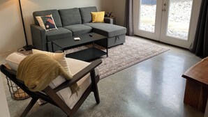 A modern living room with a grey sectional sofa, a small black coffee table, a wooden side table, and a floor lamp. - Lovely 2-bedroom cottage located in downtown Mount Vernon! (Mount Vernon)