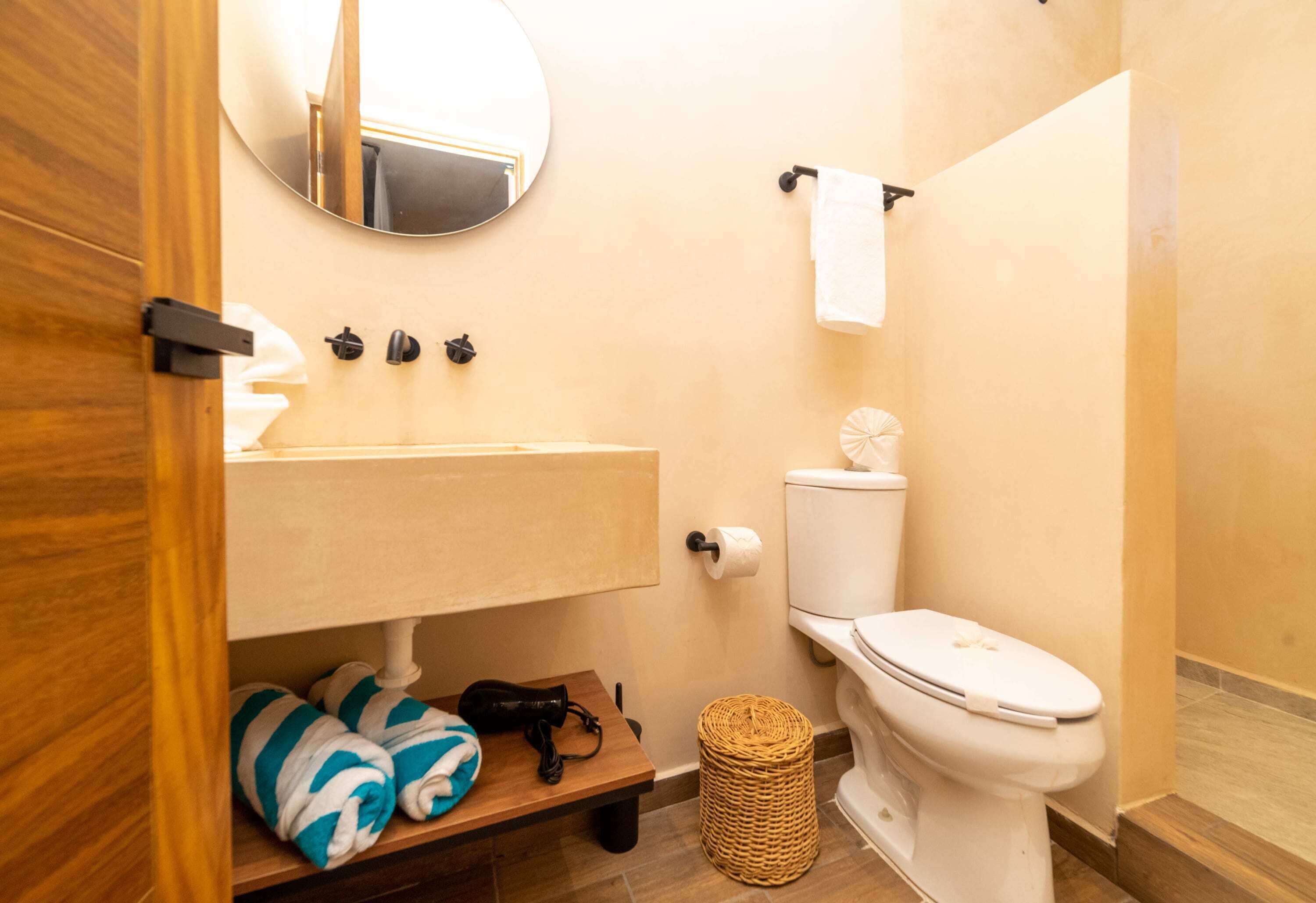 Ein Badezimmer mit einem Holzwaschtisch, einer Toilette, einem runden Spiegel und einem Korbwäscheschrank.
