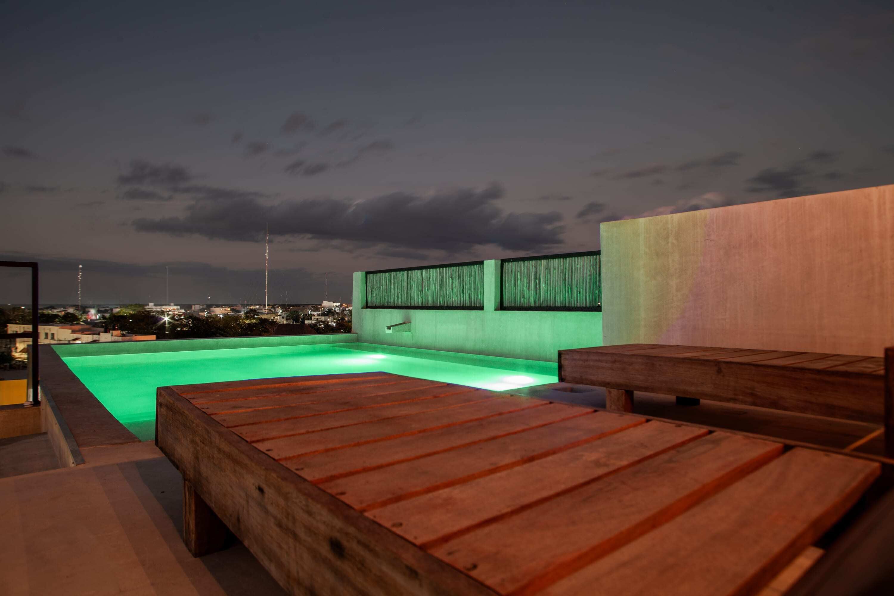 A rooftop pool area with wooden loungers and a green-lit pool.