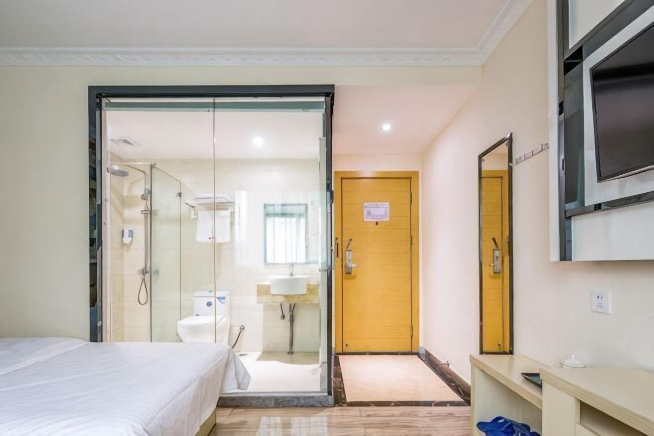 Habitación de hotel con cama, baño con ducha, lavamanos y espejo. Hay una puerta que conduce a otra habitación y un televisor montado en la pared.