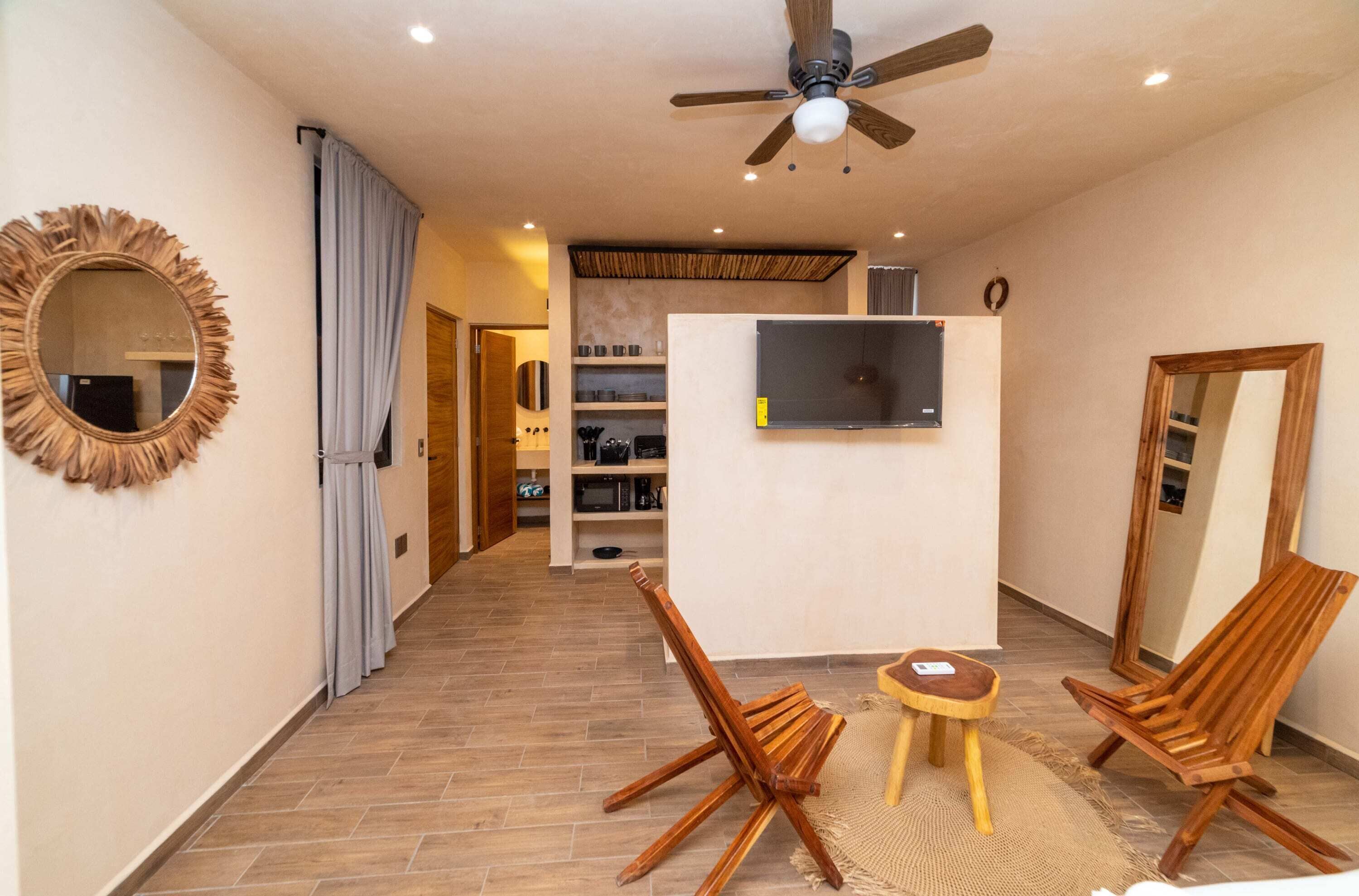 Un salon moderne doté d’un ventilateur de plafond, d’un téléviseur fixé au mur, de chaises en bois, d’une petite table ronde et d’un grand miroir.