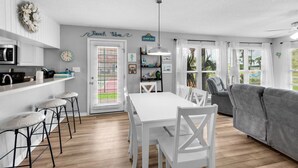 A modern kitchen with white cabinets, a dining area with a white table and chairs, and a gray sofa in a bright living room. - 🌊Beachview 107-🐾Pet Friendly🐾-2BR-Close To The Beach🌊 (Navarre Beach)