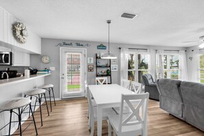 A modern kitchen with white cabinets, a dining area with a white table and chairs, and a gray sofa in a bright living room. - 🌊Beachview 107-🐾Pet Friendly🐾-2BR-Close To The Beach🌊 (Navarre Beach)