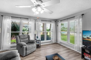 A spacious living room with a ceiling fan, large windows with blinds, a grey sofa, a small table with a lamp, and a TV on a stand.