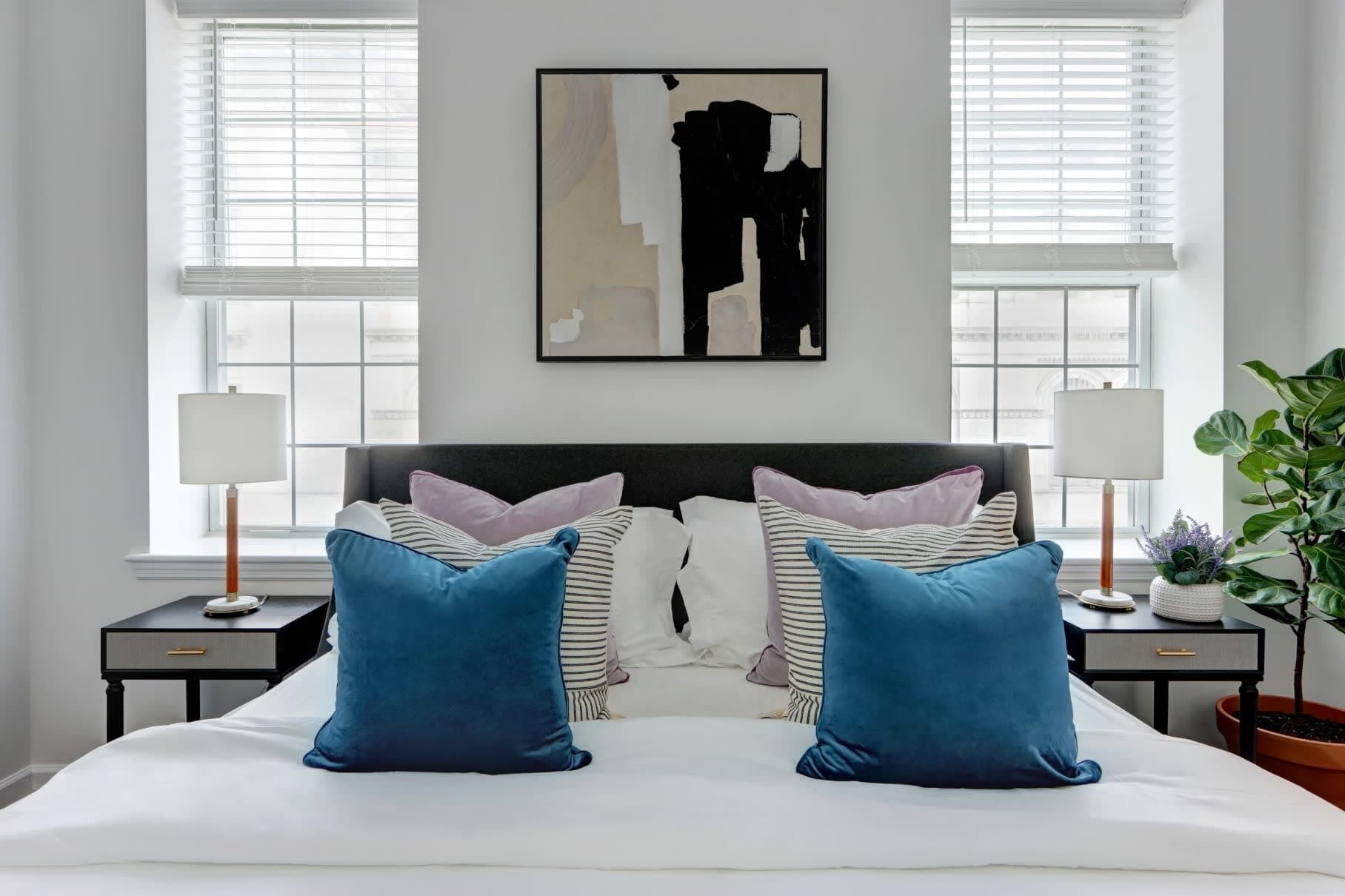 Ein Schlafzimmer mit einem großen Bett, zwei Nachttischen mit Lampen, einem Wandbild im abstrakten Stil und einer Zimmerpflanze.
