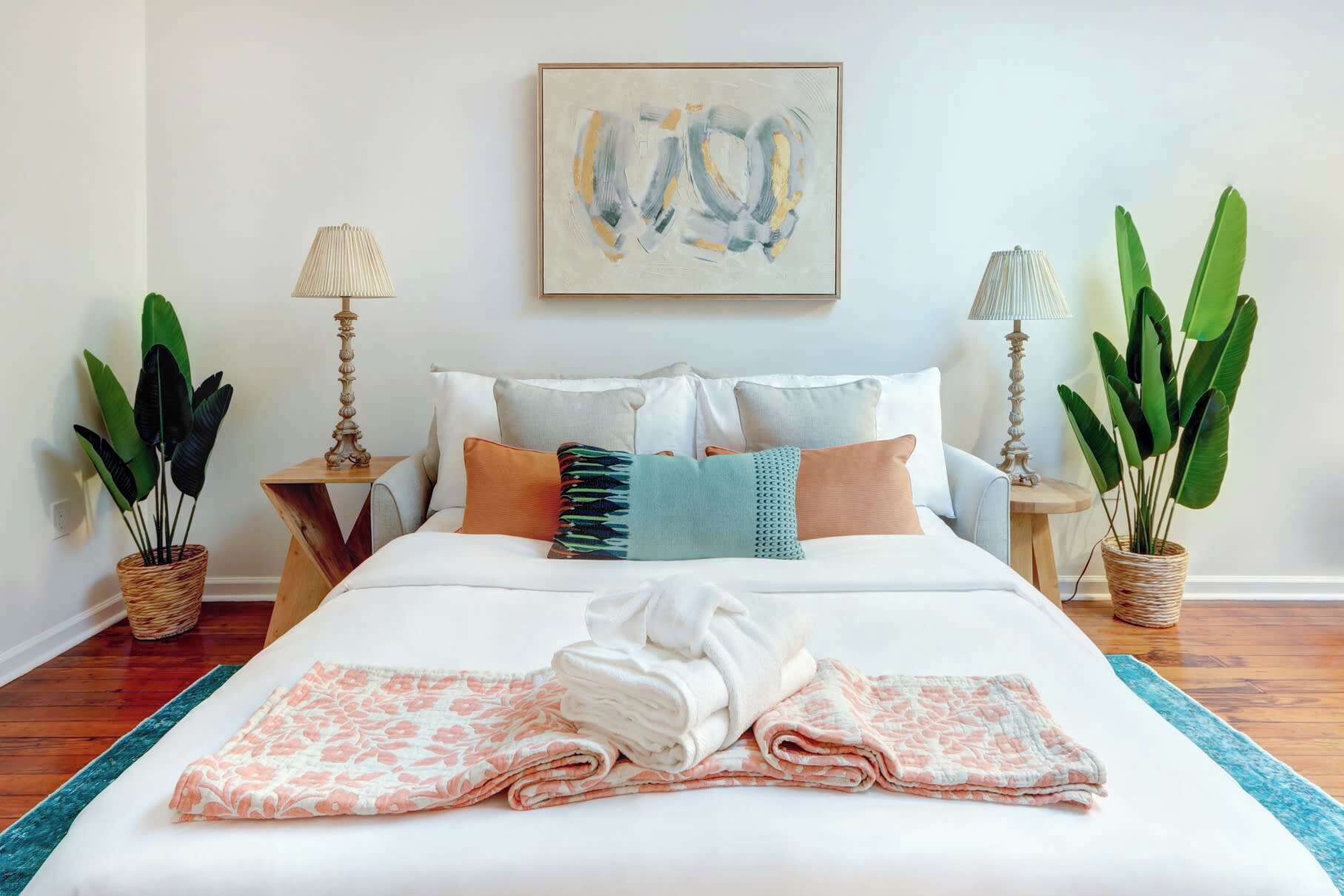 A neatly made bed with pillows, a blanket, and a folded towel. There are two bedside tables with lamps and potted plants.