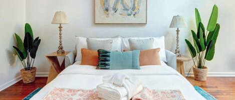 A neatly made bed with pillows, a blanket, and a folded towel. There are two bedside tables with lamps and potted plants.