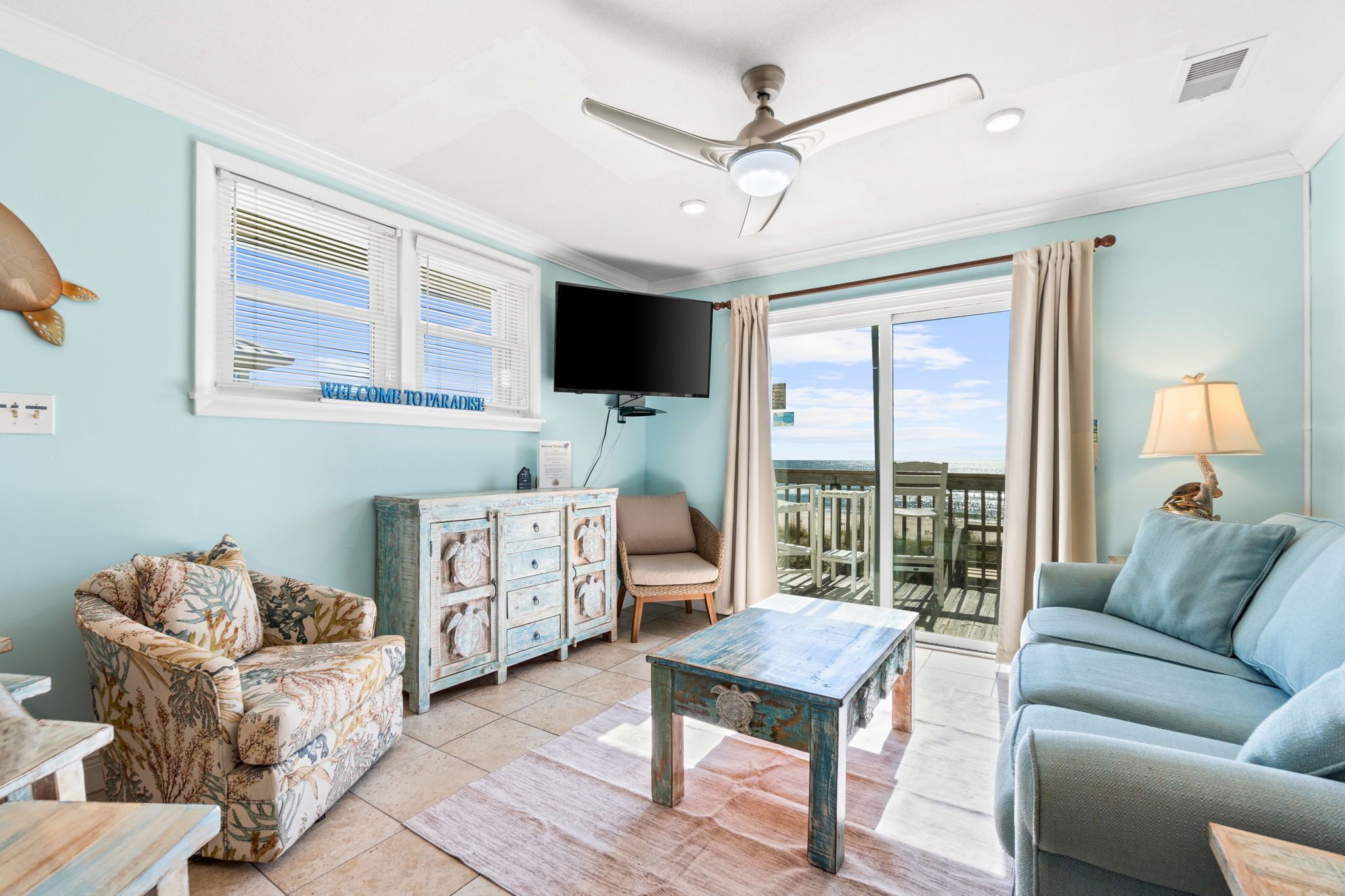 A living room with a ceiling fan, a television, a sofa, a chair, a coffee table, and a view of the outdoors.