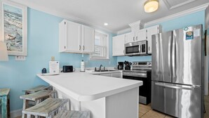 Eine moderne Küche mit weißen Schränken, einem Edelstahlkühlschrank, einer Mikrowelle und einem Backofen. Eine Holzeinsel bietet Sitzgelegenheiten und an der Wand hängt ein gerahmtes Bild.