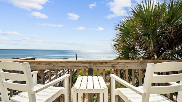 Eine Holzterrasse mit weißen Stühlen mit Blick auf einen Strand.