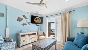 Ein Wohnzimmer mit blauem Sofa, einem Holztisch, einem an der Wand montierten Fernseher und Blick auf das Meer durch eine Glastür.