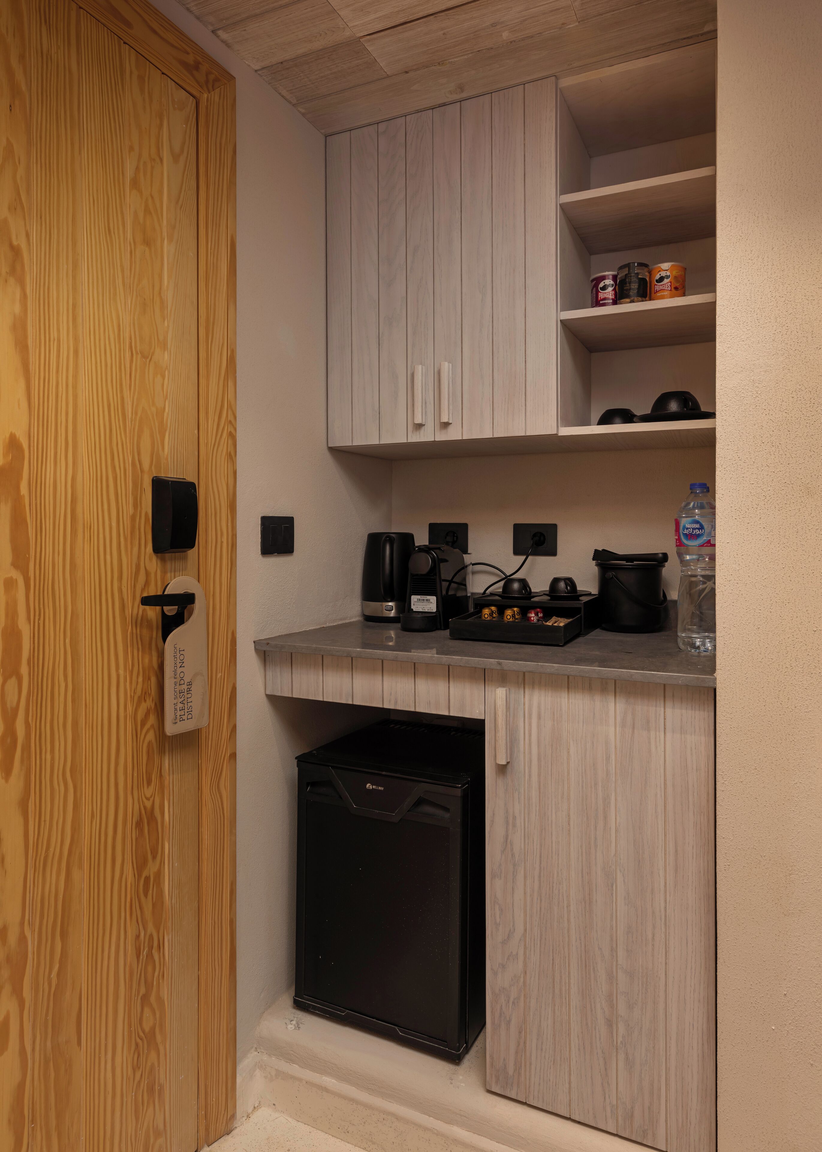 A compact kitchenette with a microwave, coffee maker, and small refrigerator. There are wooden cabinets above the counter and a door with a "Do Not Disturb" sign.