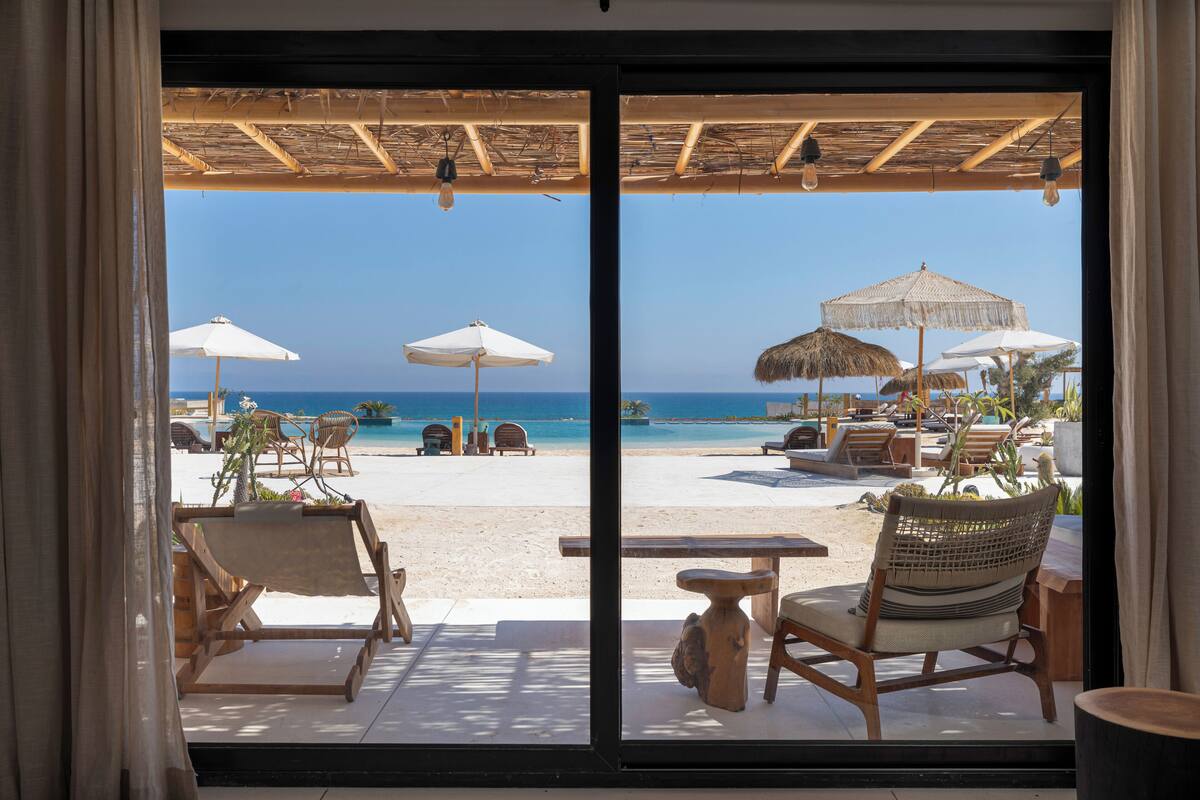 Une chambre avec de grandes fenêtres offrant une vue sur la plage, et dotée d’un espace extérieur aménagé avec des parasols et des chaises longues.