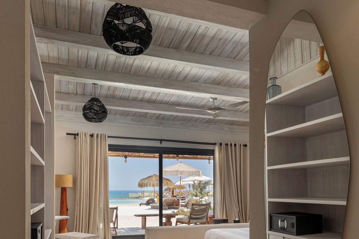 Quarto com uma grande janela, uma cama, um banco de madeira e vista para a praia.