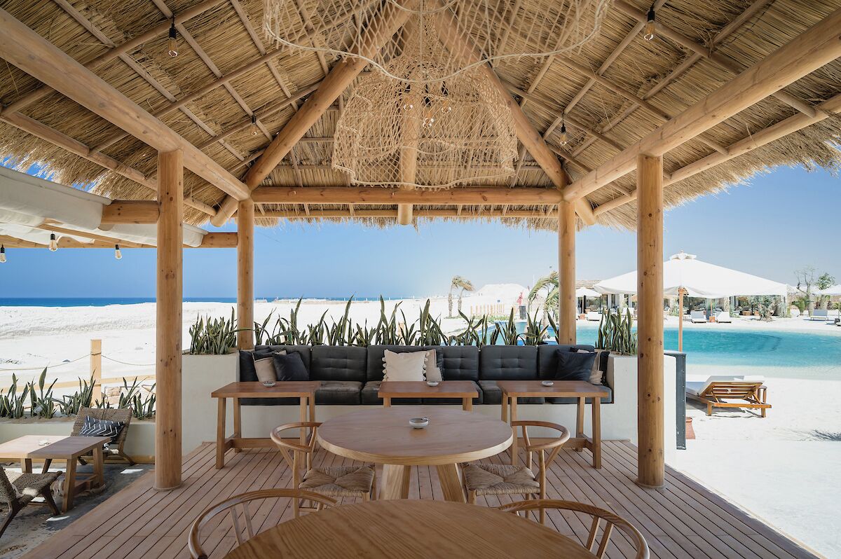 Un área de descanso junto a la playa, con techo de paja, mobiliario de madera y vista al océano y la playa.