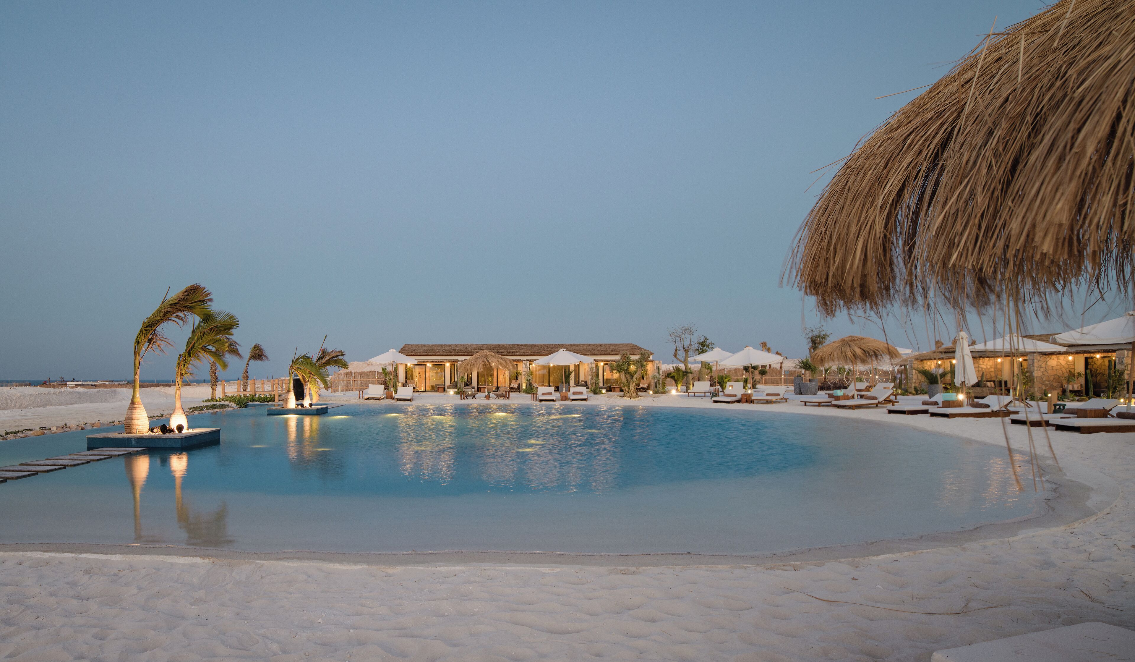 A resort pool with sun loungers and umbrellas, surrounded by a sandy beach and palm trees.