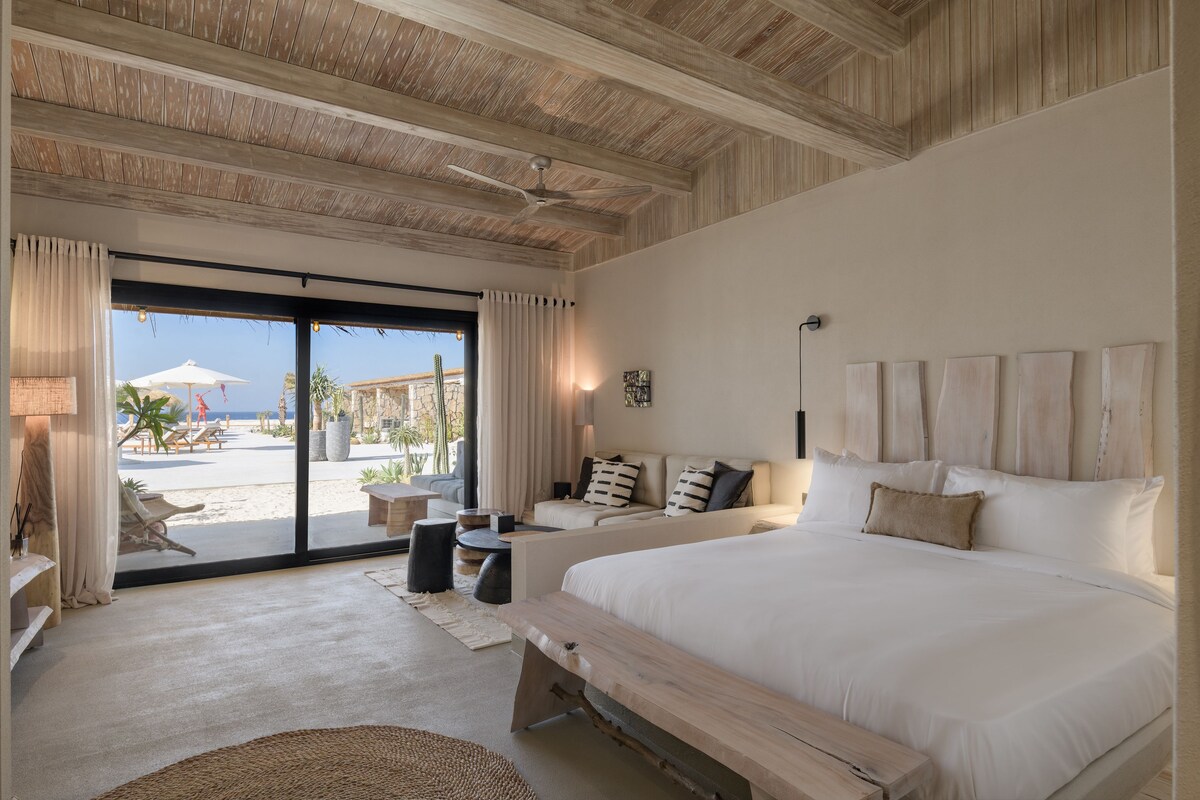 Um quarto espaçoso com uma cama grande, um sofá e um banquinho. Possui uma porta de vidro deslizante com vista para a praia.