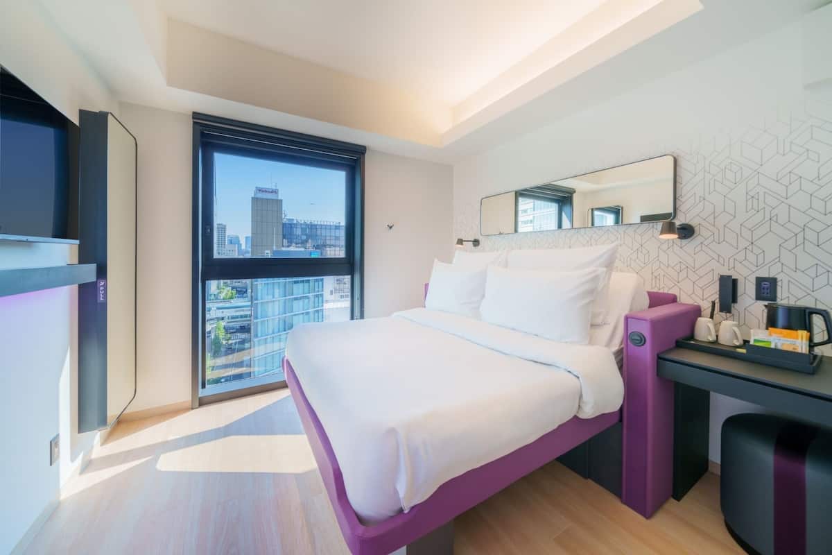 A modern hotel room with a large bed, a desk, a mirror, and a city view.
