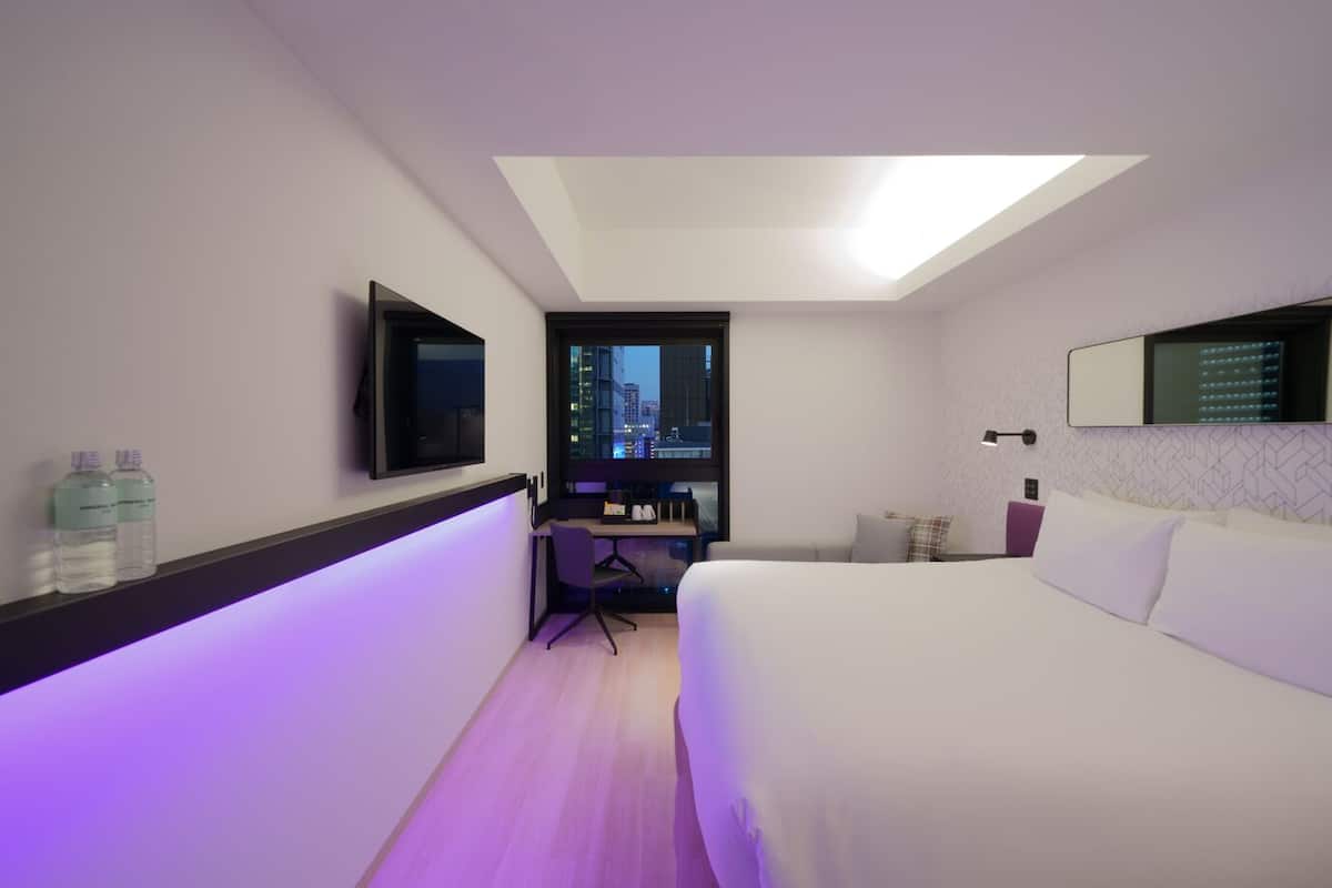 A modern hotel room with a bed, desk, chair, and a large window with a city view.