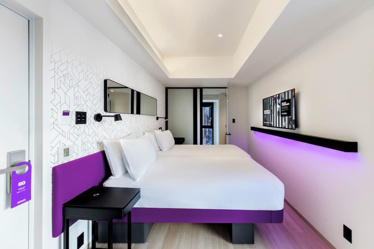 A modern hotel room with a large bed, a bedside table, a mirror, and a wall-mounted shelf.