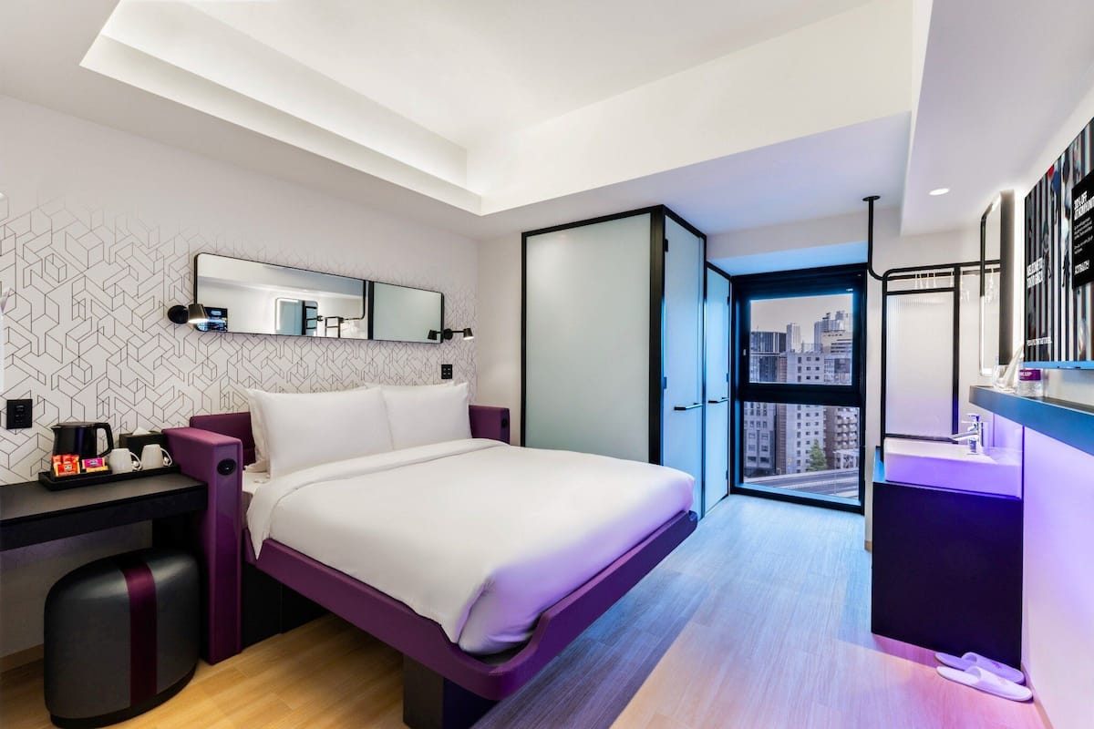 A modern hotel room with a bed, a desk, a chair, a small table with a coffee maker, a mirror, and a view of the city through a large window.