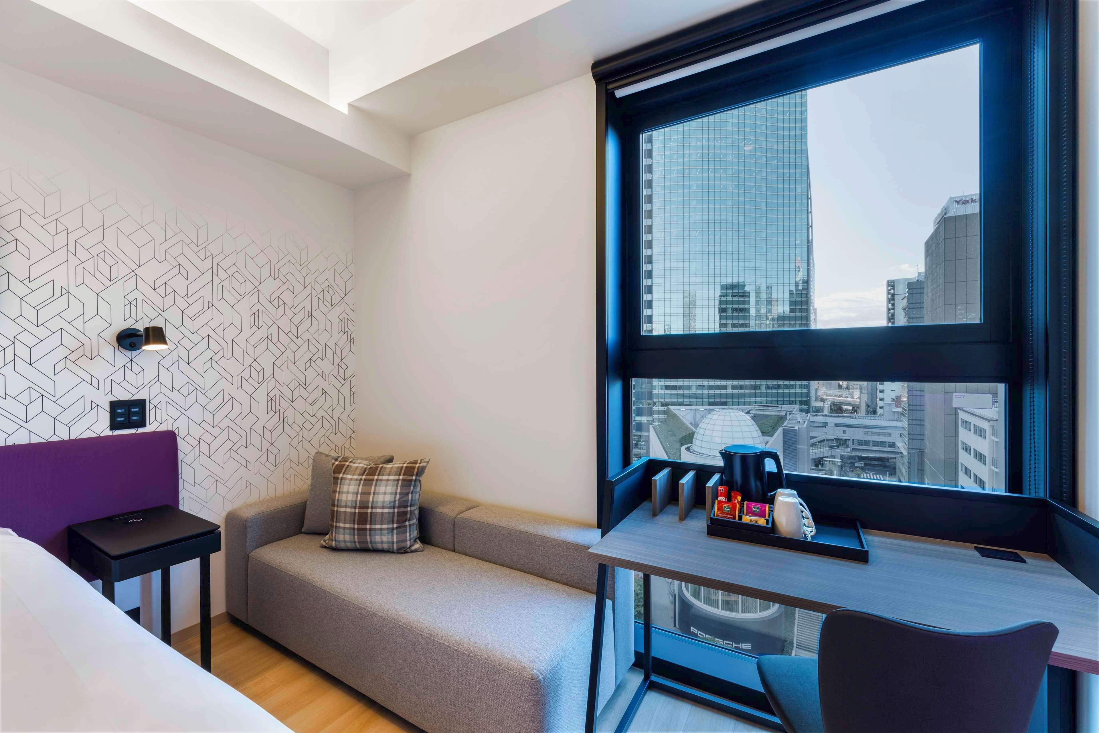 A modern hotel room with a sofa, a small table with a coffee maker, and a large window offering a city view.