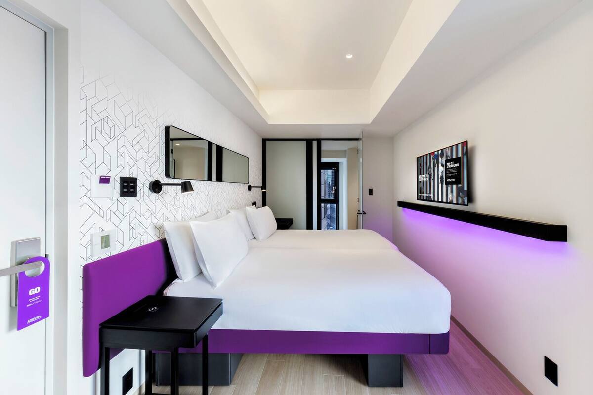 A modern hotel room with a large bed, a bedside table, a wall-mounted TV, and a door with a purple sign.
