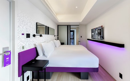 A modern hotel room with a large bed, a bedside table, a wall-mounted TV, and a door with a purple sign.