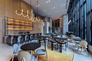 Zona de bar moderna con una barra larga, varios taburetes y mesas. Amplias ventanas a un lado y un área de estar con mesas redondas y sillas.