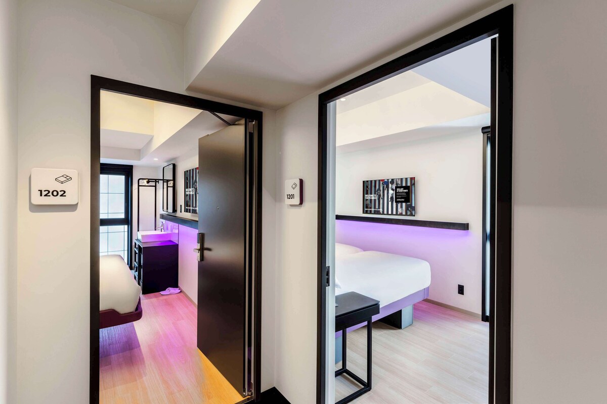 A modern hotel room with a bed, bedside table, and a small desk. There is a window with a view of the city.