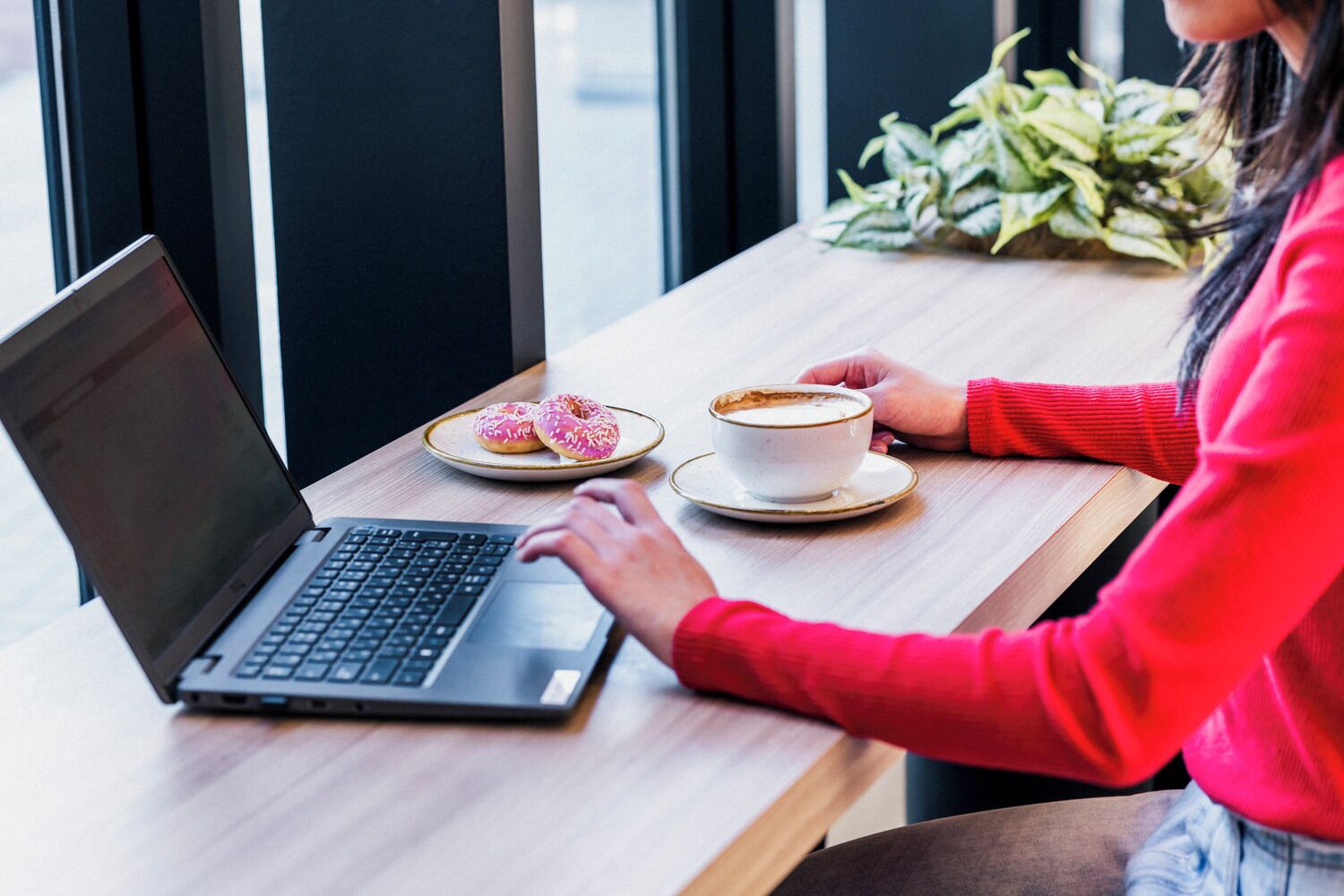 A person using a laptop at a wooden table with a cup of coffee and two donuts.